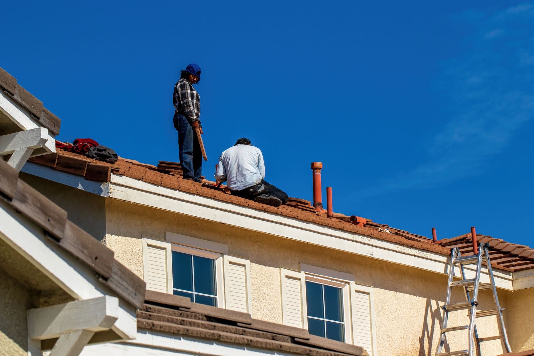 Two workers repairing tiles on a tiled roof, ladder at the eaves